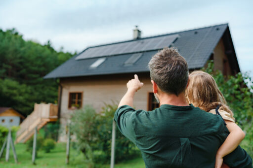 Rear View Of Dad Holding Her Little Girl In Arms And Showing At Their House With Installed Solar Panels. Alternative Energy, Saving Resources And Sustainable Lifestyle Concept.